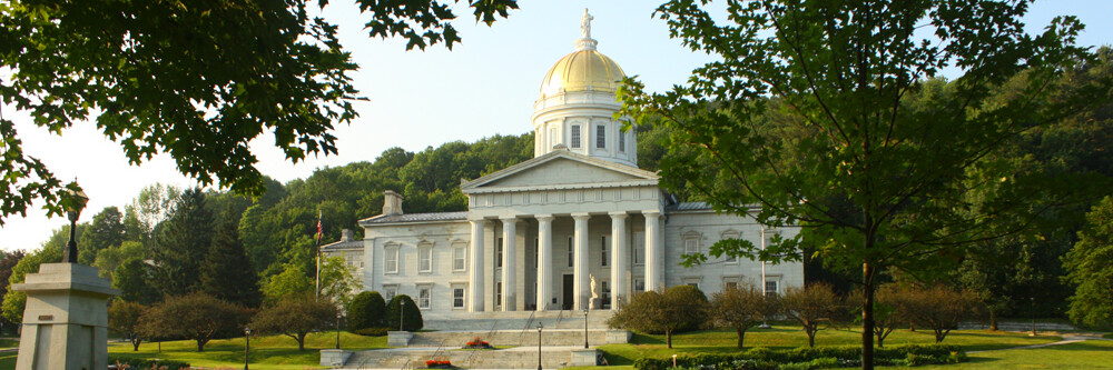 Vermont State House with a golden dome, surrounded by trees and green landscape.